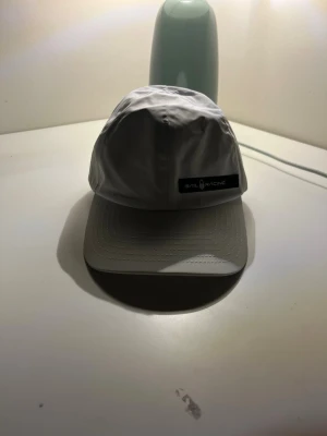 Vit keps från Sail Racing - Vit keps från Sail Racing med böjd skärm och justerbar rem baktill. Kepsen har en stilren design med logotyp på framsidan och är tillverkad i ett lätt syntetmaterial. Perfekt för dig som gillar sportig och clean stil.