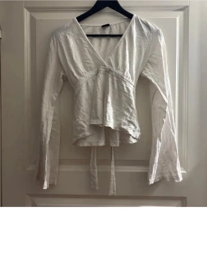 Vit omlottblus med knytband - Säljer en vit omlottblus med långa ärmar och knytband i midjan. Blusen har en djup v-ringning och är tillverkad i ett lätt tyg som ger en avslappnad look. Perfekt för att skapa en trendig och stilren outfit.
