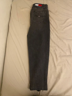 Svarta mom jeans från Tommy Jeans - Säljer ett par svarta mom jeans från Tommy Jeans med hög midja och raka ben. Klassisk femficksmodell med logga på bakfickan och bältesslingor. Jeansen är gjorda i kraftig denim och har en snygg vintage vibe.