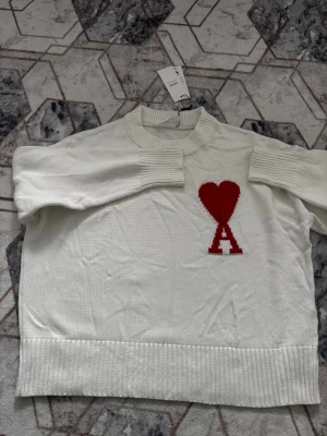 Vit stickad tröja från Ami Paris. Helt ny - Vit stickad tröja från Ami Paris med röd hjärtlogga och bokstaven A på bröstet. Tröjan har ribbade muddar vid ärmslut, hals och nederkant samt en avslappnad passform. Perfekt för dig som gillar stilrena plagg med en twist. Helt ny och priset kan diskuteras!