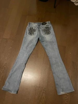 Ljusblå bootcut jeans med detaljer - Säljer ett par ljusblå bootcut jeans med snygga broderade bakfickor och nitar. Jeansen har en klassisk femficksmodell och är tillverkade i denim med lätt slitning framtill. Perfekta för dig som gillar en unik och trendig look. Innebenslängden är 82cm, rakt över midjan är 40 och rakt över låren är 23cm. De finns uppe på vinted också om du har lättare o köpa där (idakeisuu)