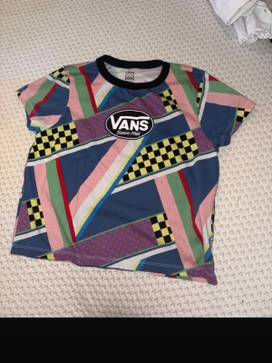 Färgglad mönstrad Vans t-shirt - Säljer en riktigt färgstark och unik t-shirt från Vans med coola grafiska mönster i blått, rosa, gult, grönt och svart. T-shirten har korta ärmar, rund hals och det klassiska Vans-loggan framtill. Perfekt för dig som vill sticka ut med din stil.