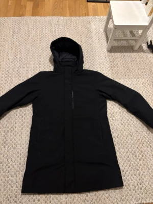 Svart lång parkas från Uniqlo - Stilren svart parkas från Uniqlo med huva och dold dragkedja. Jackan är vadlång och har två sidofickor samt quiltat innerfoder för extra värme. Perfekt för kalla dagar och enkel att matcha med olika outfits.