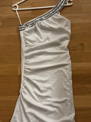Vit oneshoulder klänning med paljetter. - Snygg vit oneshoulder klänning med silverfärgade paljettdetaljer längs axelbandet. Klänningen har en figurnära passformen .Perfekt för dig som vill sticka ut på nästa event.