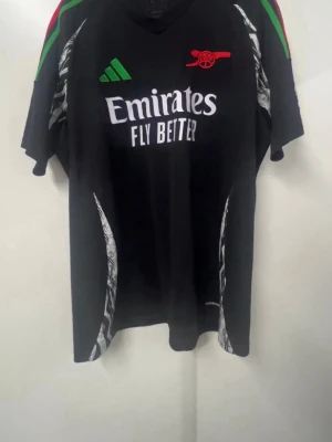 Arsenal Adidas svart matchtröja XL - Snygg svart Arsenal matchtröja från Adidas med gröna och röda detaljer på axlarna, vita mönster längs sidorna och klubbens logga i rött. Tröjan har 'Emirates Fly Better' tryckt på bröstet och är i slim fit-modell. Perfekt för fotboll eller träning.