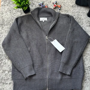 Grå stickad ziptröja Maison Margiela - Snygg grå stickad ziptröja från Maison Margiela med hög krage och hel dragkedja framtill. Tröjan har ribbstickade muddar och en klassisk, avslappnad passform. Perfekt för lager-på-lager och stilrena outfits.