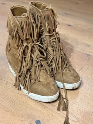 Bruna mocka boots med fransar - säljer ett par bruna boots i mocka med coola fransdetaljer och snörning framtill. Skorna har vit sula och en unik design som sticker ut. Perfekta för dig som vill ha en bohemisk och trendig look. Använda några gånger men måste sälja de är inte min storlek!