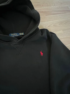 Svart hoodie från Polo Ralph Lauren - Svart hoodie från Polo Ralph Lauren med klassisk huva och röd broderad logga på bröstet. Tillverkad i mjukt bomullsmaterial som känns skönt mot huden. Perfekt för en avslappnad och stilren look. Lite lång för mig som är 161 cm XL i barnstorlek. 