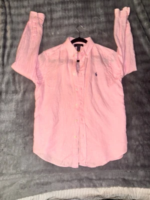 Rosa linneskjorta från Ralph Lauren - Säljer en ljusrosa skjorta från Ralph Lauren i linne med klassisk button-down krage och broderad logga på bröstet. Skjortan har lång ärm och knappar framtill. Perfekt för en avslappnad men ändå stilren look.