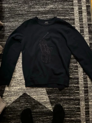 Sweatshirt från Polo Ralph Lauren - Svart sweatshirt med rund hals från Polo Ralph Lauren. Stor broderad logga med polospelare framtill i samma svarta ton som tröjan. Mjuk och bekväm, perfekt till jeans eller joggers.