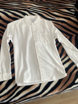 Vit skjorta från H&M med bröstficka - Säljer en vit skjorta från H&M med klassisk krage och bröstficka. Skjortan har långärmad design, knappar framtill och är tillverkad i ett strukturerat bomullsmaterial som ger en snygg och modern look. Perfekt för dig som gillar stilrena plagg.