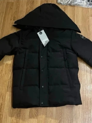 Svart pufferjacka från Canada Goose - Säljer en svart pufferjacka från Canada Goose med stor huva och patch på ärmen. Jackan har en boxig passform, stora framfickor och stängs med tryckknappar. Perfekt för kalla vinterdagar och riktigt snygg streetstil.