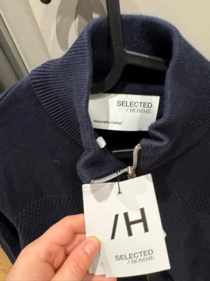 Marinblå ziptröja Selected Homme - Snygg marinblå ziptröja från Selected Homme med hög krage och strukturerat mönster över bröstet och armarna. Tröjan har långa ärmar och dragkedja framtill. Perfekt för en stilren och avslappnad look.
