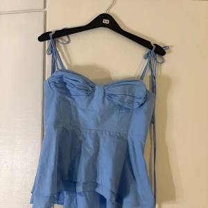 Ljusblå linnetopp med knytband - Supersöt ljusblå linnetopp med tunna axelband som knyts på axlarna. Toppen har en söt volang nedtill och veckade detaljer vid bysten. Perfekt för varma dagar och ger en romantisk vibe till din outfit.