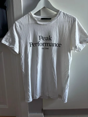 Vit Peak Performance t-shirt - Klassisk vit t-shirt från Peak Performance med svart tryck på bröstet. T-shirten har rund hals och korta ärmar, perfekt för en clean och enkel stil. Tillverkad i mjuk bomull som känns skön mot huden.