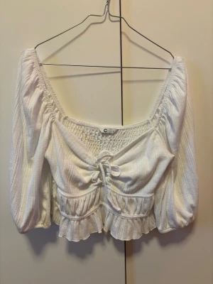 Vit croppad blus med snörning stl S - Supersöt vit croppad blus från Cubus med puffiga trekvartsärmar, snörning framtill och volangkant nedtill. Toppen har ribbad struktur och smockad rygg för extra bekväm passform. Perfekt för dig som gillar romantisk och trendig stil. Tvättad men aldrig använd. 