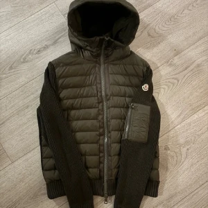 Mörkgrön cardigan från Moncler - Snygg mörkgrön cardigan från Moncler med stickade ärmar och huva. Jackan har en quiltad kropp, dragkedja framtill och en ficka med dragkedja på ena ärmen. Moncler-logga på ärmen ger en exklusiv touch. Perfekt för kalla dagar.