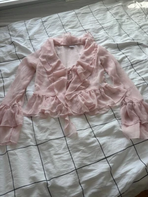 Ljusrosa volangblus med knytband - Supersöt ljusrosa blus med volanger och knytband framtill. Blusen har långa ärmar med volangdetaljer och är gjord i ett tunt, skirt tyg som ger en romantisk känsla. Perfekt för dig som gillar en feminin och trendig stil.