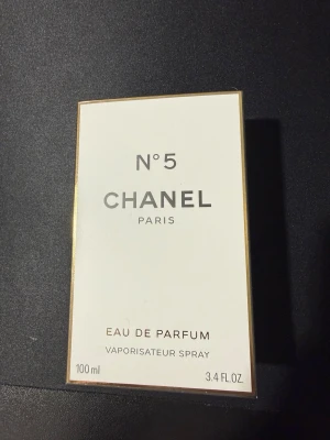 Chanel N°5 Eau de Parfum 100 ml - Chanel N°5 Eau de Parfum i klassisk vit och guldig kartong. Flaskan innehåller 100 ml och är en ikonisk doft från Chanel Paris. Förpackningen har stilren design med svarta och guldiga detaljer. Perfekt för dig som älskar exklusiva och tidlösa parfymer.