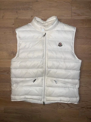Vit Moncler Gui väst - Vit Moncler Gui väst i size 2. med hög krage och två dragkedjeförsedda fickor framtill. Västen har quiltad design och klassisk Moncler-logga på bröstet. Perfekt för lager-på-lager och ger en clean look till din outfit.
