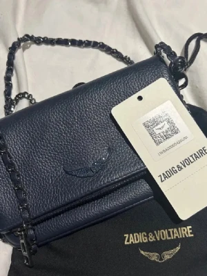 Mörkblå handväska Zadig & Voltaire - Snygg mörkblå handväska från Zadig & Voltaire i strukturerat läder. Väskan har en kedjerem och ett emblem med vingar på framsidan. Perfekt för dig som vill ha en stilren och trendig accessoar med coola detaljer.