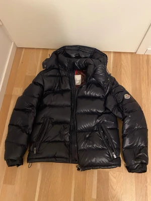 Svart dunjacka från Moncler - Snygg svart dunjacka från Moncler med glansig finish och klassisk logga på ärmen. Jackan har två dragkedjeförsedda fickor fram, hög krage och justerbar huva. Perfekt för kalla vinterdagar och riktigt trendig look.