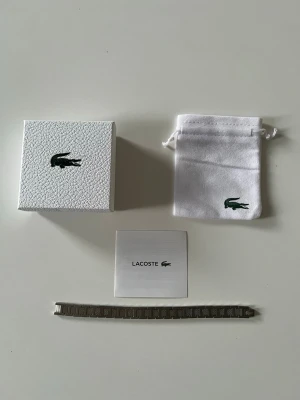 Silverfärgat armband från Lacoste - Stilrent armband i silverfärgad metall från Lacoste med rektangulära länkar och diskret Lacoste-logga. Kommer med original ask och tygpåse med krokodiltryck. Perfekt accessoar för en clean och modern look. Cirka 20 cm oanvänd har en långbortagare som kan anpassa till din storlek 