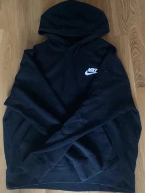 Svart Nike hoodie med logga - Säljer en svart hoodie från Nike med klassisk vit logga på bröstet. Hoodien har huva och en stor magficka framtill. Tillverkad i mjukt material som är skönt att bära. Perfekt för en avslappnad och sportig stil.