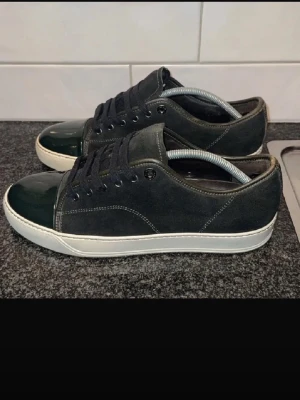 Svarta sneakers från Lanvin - Snygga svarta sneakers från Lanvin med ovandel i mocka och tåparti i blankt läder. Skorna har svarta skosnören, vita sulor och metallfärgade öljetter. Perfekta för dig som gillar stilrena och exklusiva sneakers.