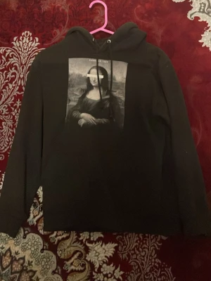 Svart hoodie med Mona Lisa-tryck - Cool svart hoodie med stort Mona Lisa-tryck på bröstet där ögonen är censurerade med ett vitt streck. Texten 'Florence, 1503' syns ovanför motivet. Hoodien har huva med dragsko och långa ärmar. Perfekt för dig som gillar streetwear och konstinspirerade plagg.