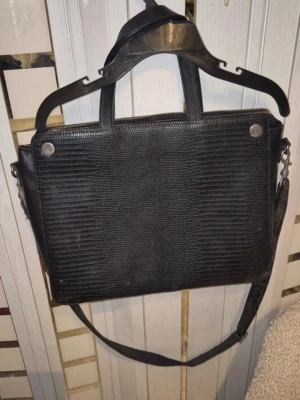 Svart handväska i ormmönstrat skinn - Snygg svart datorväska med ormmönstrad yta och både handtag och justerbar axelrem. Väskan har en stor ytterficka med dragkedja och silverfärgade detaljer. Perfekt för laptop och viktiga prylar. Stilren och passar till det mesta. Märket är Åhléns 