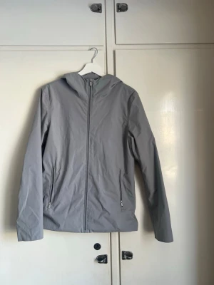 Grå vindjacka från elvine  - Snygg grå vindjacka med huva och dragkedja framtill. Jackan har två praktiska fickor med dragkedja och är tillverkad i ett lätt och smidigt material som passar perfekt när det blåser. Enkel och stilren design som är lätt att matcha.