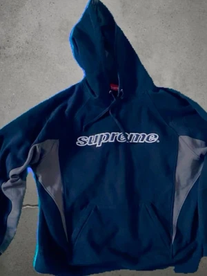 Supreme svart hoodie med grå detaljer - Supreme hoodie i svart med grå paneler på sidorna och ärmarna. Stor Supreme-logga broderad på bröstet, siffran 24 på ryggen och texten 'Division One Champs' längst ner bak. Klassisk känguruficka och huva med dragsko. Perfekt för streetwear-stilen.
