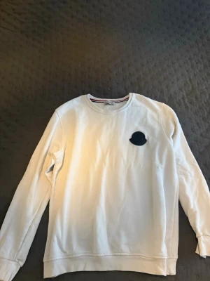 Vit sweatshirt från Moncler - Snygg vit sweatshirt från Moncler med rund halsringning och svart Moncler-logga på bröstet. Tröjan har en klassisk passform och långa ärmar. Perfekt för en clean och stilren look.