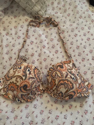 Mönstrad bikiniöverdel med knyt - Snygg bikiniöverdel med paisley-inspirerat mönster i beige, orange och blå toner. Halterneck-modell med knyt i nacken och justerbart spänne i ryggen. 