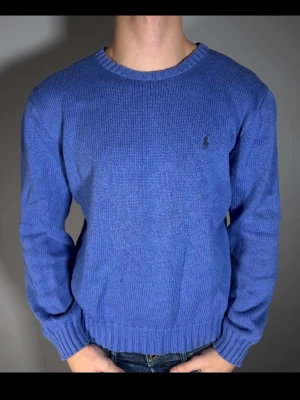 Blå stickad tröja från Ralph Lauren - Klassisk blå stickad tröja från Ralph Lauren med rund halsringning och diskret broderad logga på bröstet. Tröjan har ribbade muddar vid ärmslut och nederkant samt en normal passform. Perfekt att styla med jeans eller kjol.