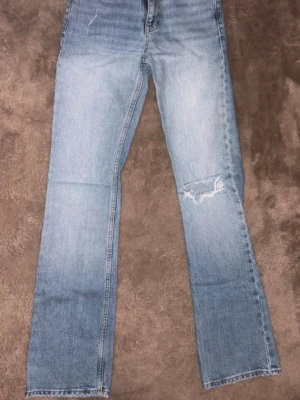 Gina tricot bootcut jeans - Säljer ett par ljusblå jeans med rak passform och en snygg slitning på ena knät. Jeansen har klassisk femficksdesign och är tillverkade i denim. Perfekta för en avslappnad och trendig look.