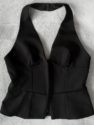 Svart halterneck korsettopp - Snygg svart korsettopp med halterneck och markerad byst. Toppen har en figurnära passform och stängs med dragkedja i sidan. Perfekt för att lyfta vilken outfit som helst.