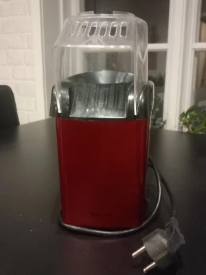 Röd popcornmaskin med transparent lock - Popcornmaskin i röd plast med svart och transparent lock. Maskinen har en elektrisk sladd och är designad för att göra popcorn snabbt och enkelt hemma. Perfekt för filmkvällar och snacks. Kompakt och lätt att använda.