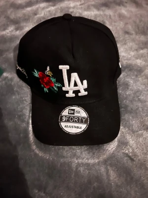 Svart LA-keps från New Era - Snygg svart keps från New Era med LA-logga broderad i vitt framtill. På sidan finns en broderad röd ros och en liten bi, vilket ger kepsen en unik touch. Modellen är 9FORTY och har justerbar rem bak för perfekt passform.