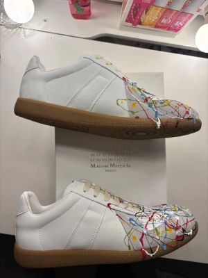 Maison Margiela vita sneakers med färgstänk - Maison Margiela sneakers i vitt läder med klassisk gummisula och unika färgstänk i rött, blått, gult och svart över tå och sidor. Låga och stilrena med en konstnärlig touch som sticker ut. Perfekta för dig som vill ha något annorlunda och trendigt.