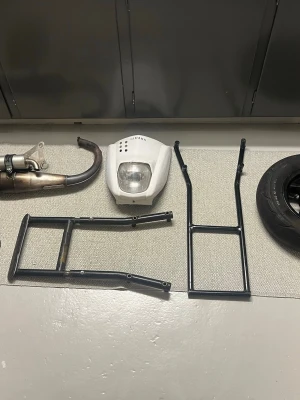 Yamaha slider delar  - Säljer ett paket med mopeddelar till Yamaha, inklusive vit frontkåpa med strålkastare, avgassystem, två svarta 12 och ett hjul med däck. Perfekt för dig som vill bygga eller laga din moped. Buda på sakerna 