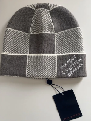 Gråvit rutig mössa  - Snygg mössa i svart och grått med rutigt mönster. Mössan är stickad och tillverkad av kashmir, ull och silke. Detalj med texten 'Marque L. Vuitton Depose' broderad framtill. Perfekt för dig som gillar exklusiva accessoarer.