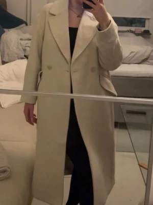Beige lång kappa från H&M - Stilren beige kappa från H&M i storlek XS. Kappan har dubbelknäppning, breda slag och två fickor med lock framtill. Lång modell som ger en klassisk och clean look. Använd 4-5 gånger max därav som ny💕💕