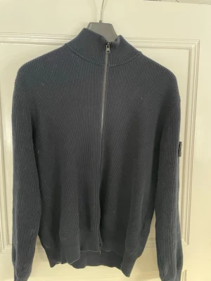 Mörkblå ziptröja från Stone Island - Mörkblå stickad ziptröja från Stone Island med hög krage och hel dragkedja framtill. Tröjan har ribbstickad struktur och den klassiska Stone Island-loggan på vänster ärm. Perfekt för lager-på-lager och snygg till jeans.   PRIS KAN DISKUTERAS 