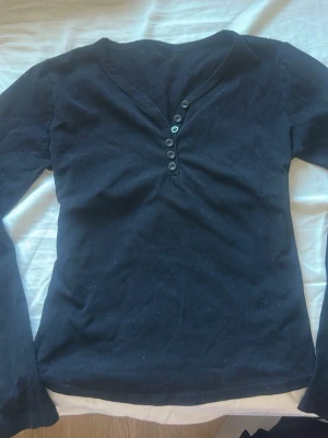 Svart långärmad topp med knappar - Snygg svart långärmad topp med v-ringning och dekorativa knappar framtill. Toppen har en enkel och stilren design som passar till det mesta. Tillverkad i mjukt bomullsmaterial för en bekväm känsla hela dagen. Det står inte riktigt storlek men den passar som en M!  endast testad!