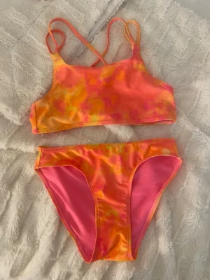 Färgglad bikini med korsade band - Snygg bikini i orange, rosa och gul med sportig topp och korsade axelband. Bikinin har ett färgstarkt mönster och klassiska bikinitrosor med rosa paneler. Perfekt för stranden eller poolen!