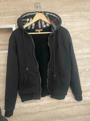 Svart hoodie från Burberry med dragkedja - Säljer en svart hoodie från Burberry med dragkedja och två fickor framtill. Huvan har det klassiska Burberry-mönstret i grått, vitt och rött. Tillverkad i mjukt bomullsmaterial och har ribbade muddar vid ärmar och nederkant. Perfekt för en stilren och avslappnad look skulle söga att den sitter mer som en S.