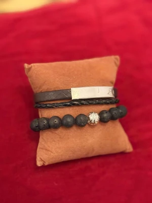 Svart armbandsset med pärlor och läder - Snyggt armbandsset med ett svart pärlarmband och ett flätat läderarmband. Pärlarmbandet har mattsvarta lavapärlor och en glittrig silverfärgad detalj i mitten. Läderarmbandet har en metallplatta och ett stilrent magnetlås i silverfärg. Perfekt för dig som gillar en modern och cool stil.