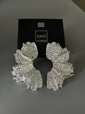 Stora silvriga örhängen SNÖ of Sweden - Unika stora örhängen från SNÖ of Sweden i silverfärgad metall. Örhängena har en rund, vågig form med en glittrig, strukturerad yta som ger ett riktigt statement-utseende. Perfekta för dig som vill sticka ut och addera något extra till din stil.